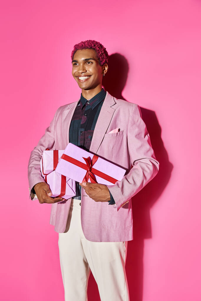 alegre hombre afroamericano en traje vibrante posando con regalos en las manos sobre fondo rosa - Foto, imagen