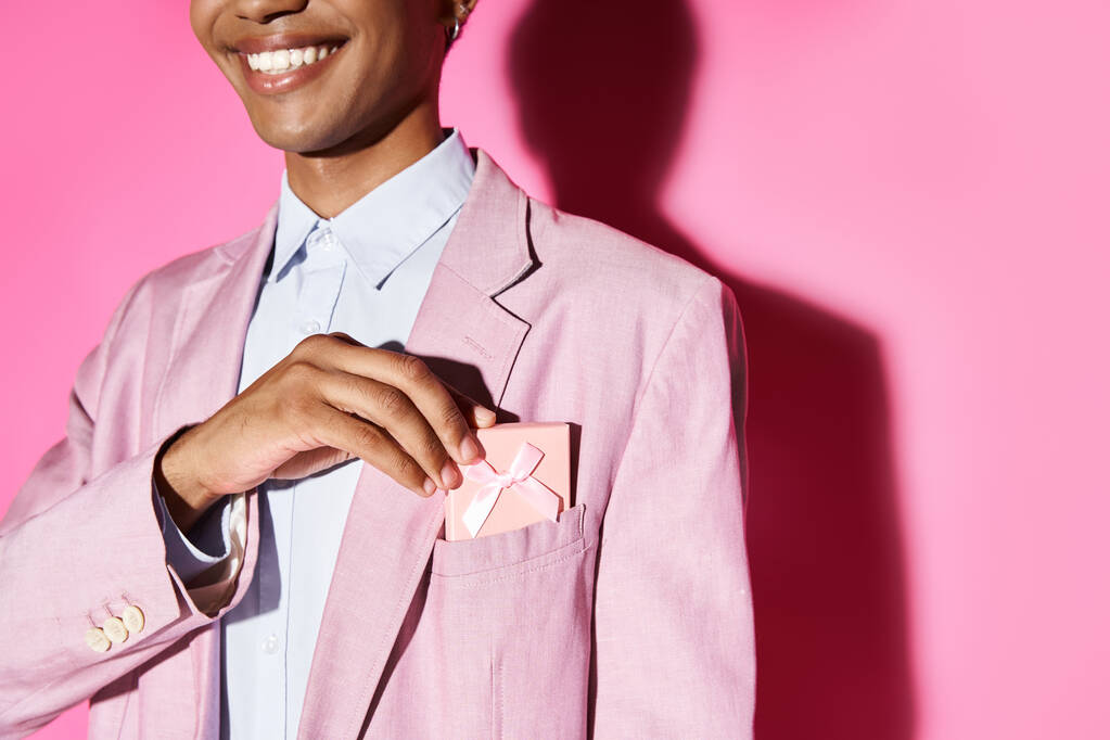 bijgesneden weergave van smiley jongeman in roze blazer zetten geschenk aan zijn zak op roze achtergrond - Foto, afbeelding