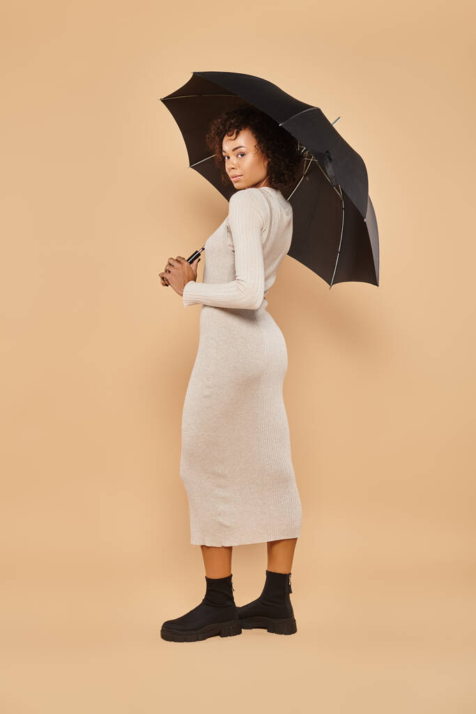 femme américaine africaine bouclée en robe midi et bottes debout sous parapluie sur beige, look automne - Photo, image