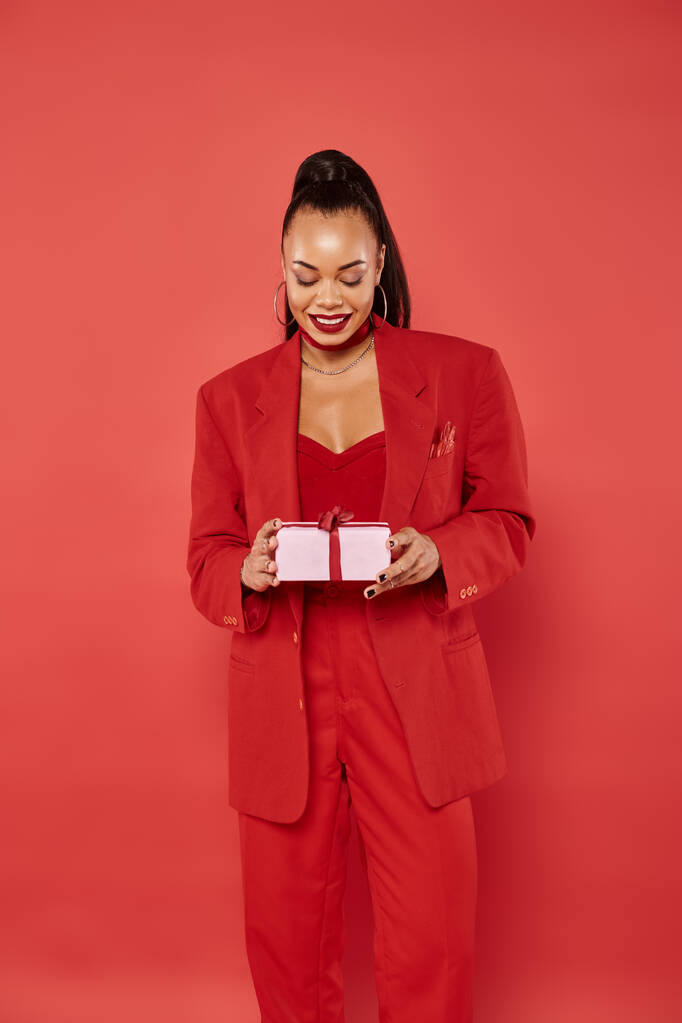 fröhliche afrikanisch-amerikanische Frau posiert im Anzug und hält Weihnachtsgeschenk auf rotem Hintergrund - Foto, Bild