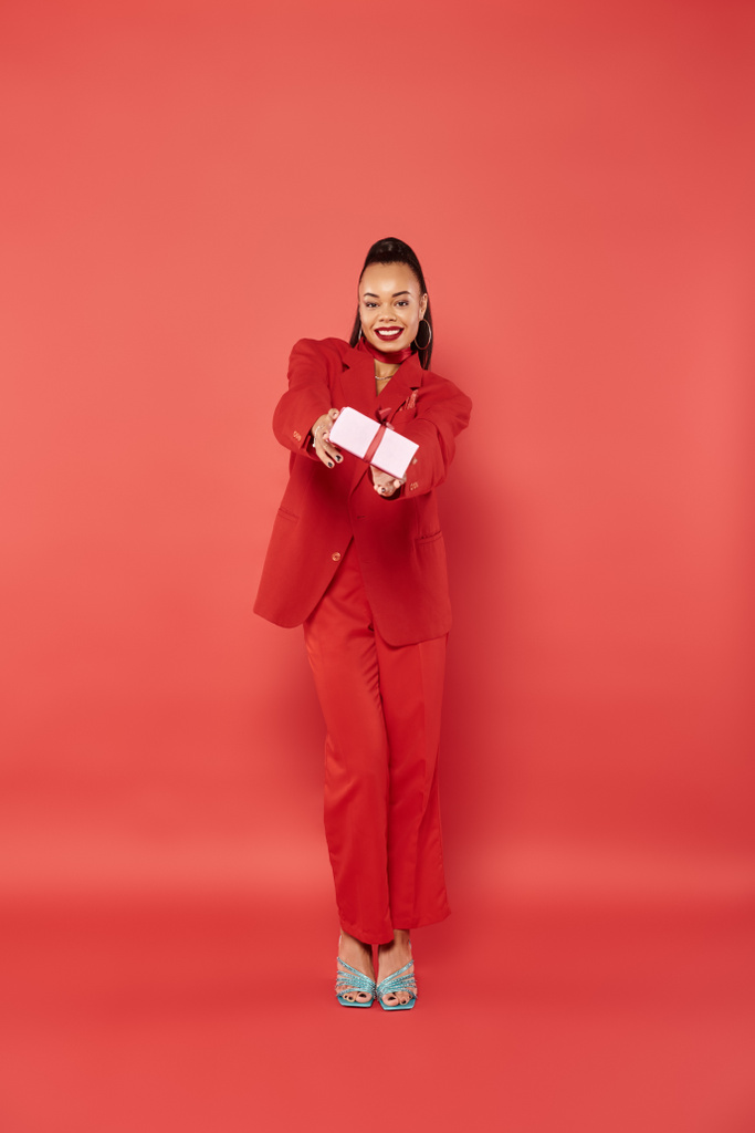 スーツを着てクリスマスプレゼントを赤い背景に持っているアフリカ系アメリカ人女性の全長 - 写真・画像