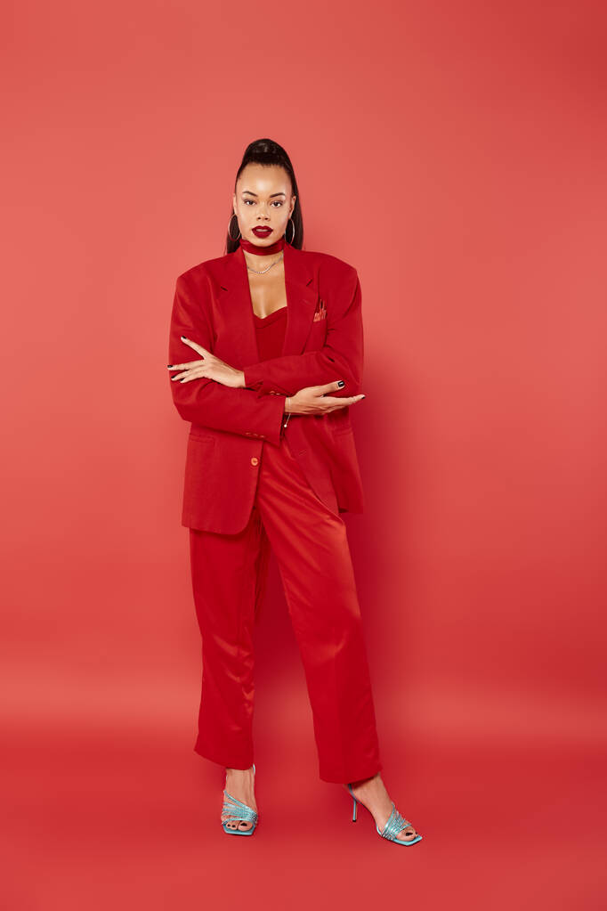 full length of beautiful african american woman with ponytail posing in suit on red background - Photo, Image
