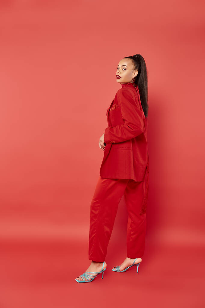 full length of beautiful african american woman with ponytail posing in suit on red background - Photo, Image