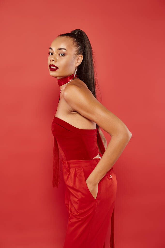 красивая африканская американская модель в красном топе без бретелек и в брюках, позирующих с рукой в кармане - Фото, изображение