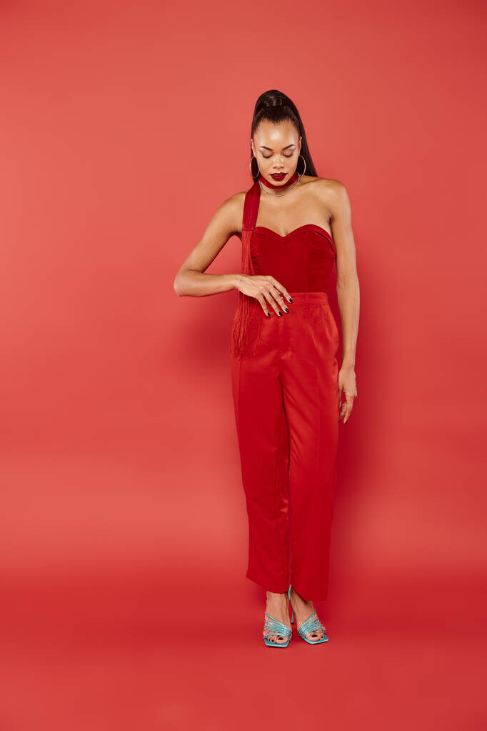 longitud completa de hermoso modelo afroamericano en top sin tirantes y pantalones posando sobre fondo rojo - Foto, imagen
