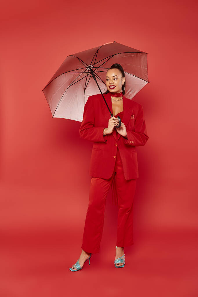 teljes hosszúságú, vidám afro-amerikai modell piros blézerben és nadrág alatt álló esernyő - Fotó, kép
