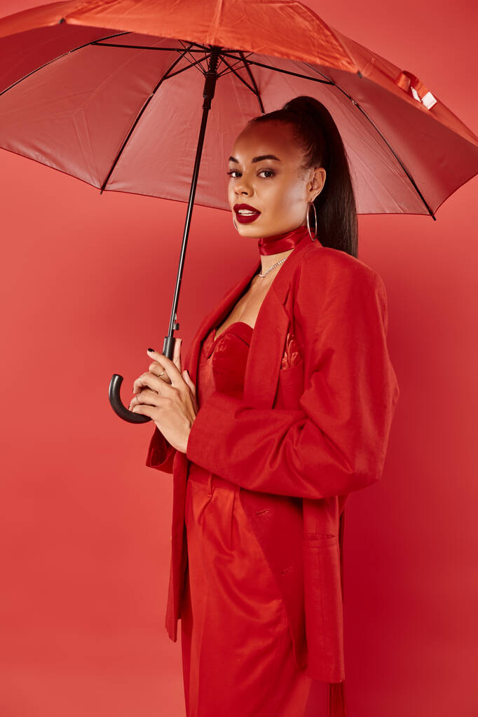 bonita mujer afroamericana en chaqueta y pantalones de pie bajo paraguas sobre fondo rojo - Foto, Imagen