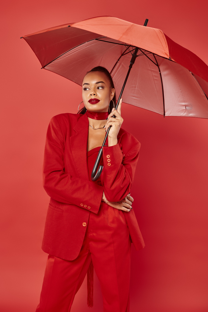 atrakcyjna młoda Afroamerykanka w kurtce i spodniach stojąca pod parasolem na czerwonym tle - Zdjęcie, obraz