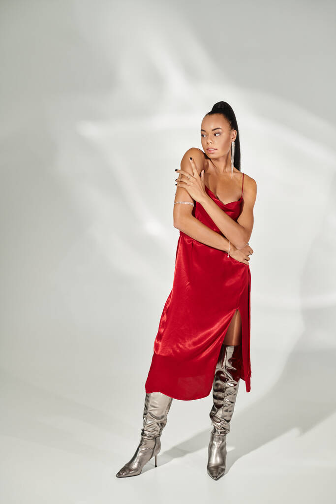 full length of brunette african american woman in red dress and silver boots on grey backdrop - Photo, Image