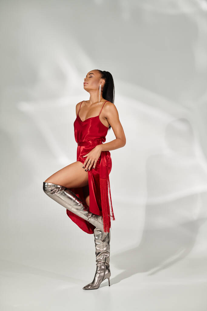 plná délka, atraktivní africká americká žena v červených šatech a stříbrné boty pózující na šedém pozadí - Fotografie, Obrázek
