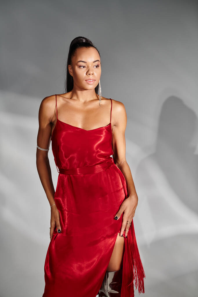 λαμπερή Αφροαμερικανή γυναίκα με κόκκινο φόρεμα κοιτάζει αλλού και ποζάρει σε καθρεφτισμένο γκρι φόντο - Φωτογραφία, εικόνα