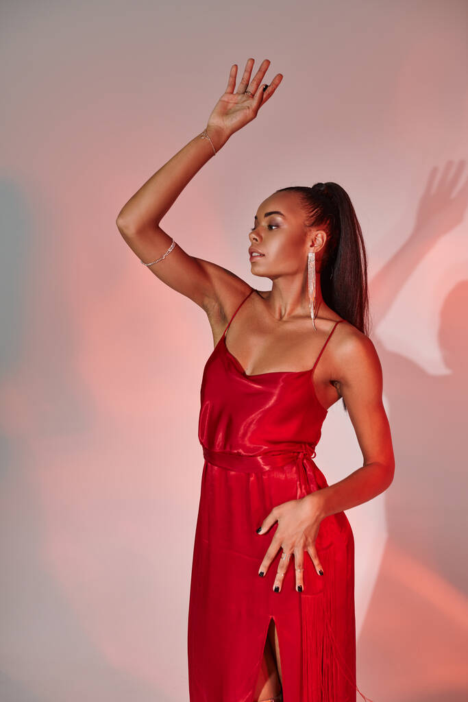 Göz kamaştırıcı Afro-Amerikalı kırmızı elbiseli kadın elini kaldırarak gri arka planda poz veriyor. - Fotoğraf, Görsel