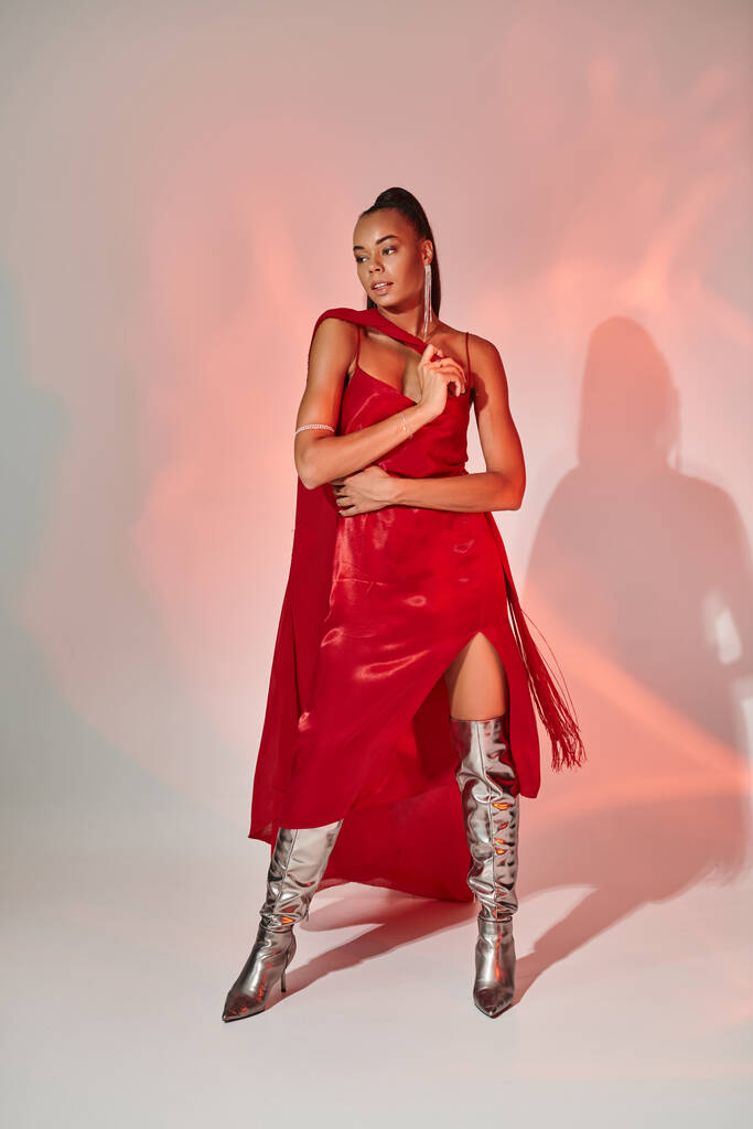όμορφη αφρικανή αμερικανίδα με κόκκινο φόρεμα με σάλι σε ασημί μπότες σε γκρι με φωτισμό - Φωτογραφία, εικόνα