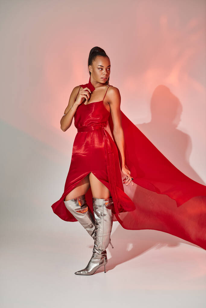 молода афроамериканка в червоній сукні з хусткою, що стоїть в срібних чоботях на сірому з освітленням - Фото, зображення