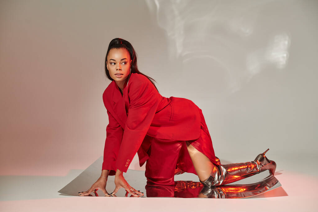 joven modelo afroamericano en blazer rojo, vestido y botas plateadas posando sobre superficie espejada - Foto, imagen