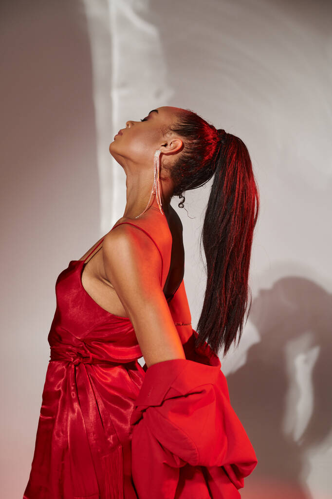 Afrikaans Amerikaans model in rode blazer en jurk poseren met gesloten ogen op grijs gespiegelde achtergrond - Foto, afbeelding