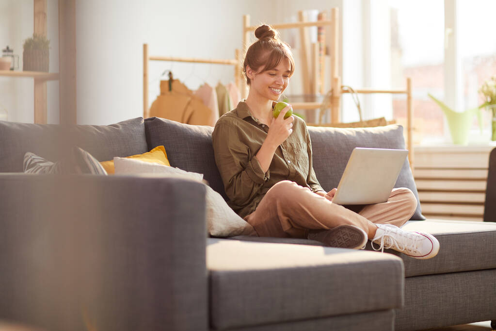 Wesoły kreatywny marketingowiec internetowy z bułką włosów siedzi ze skrzyżowanymi nogami na kanapie w sklepie i je jabłko podczas korzystania z laptopa - Zdjęcie, obraz