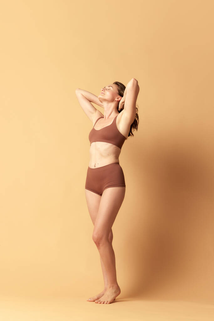 Image pleine longueur de jolie jeune femme en sous-vêtements marron debout sur fond studio beige. Concept de soins du corps et de la peau, fitness, beauté naturelle, santé, bien-être. Espace de copie pour la publicité - Photo, image