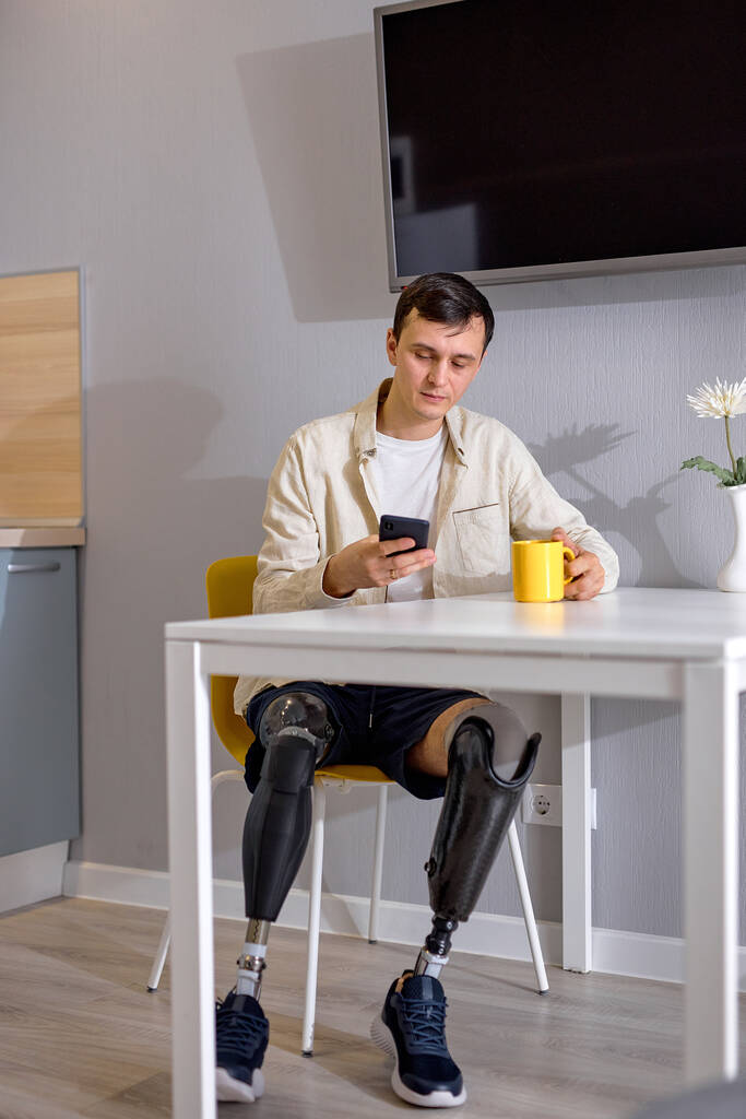 relajado chico apuesto discapacitado con biónico piernas inteligentes prótesis sentarse en la cocina, viendo la película en el teléfono inteligente y beber café o té, solo, en el interior. discapacidad física, concepto de personas - Foto, Imagen