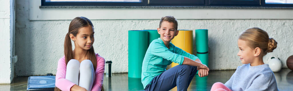 две симпатичные девочки и мальчик в спортивных костюмах сидят в спортзале и улыбаются друг другу, детский спорт - Фото, изображение