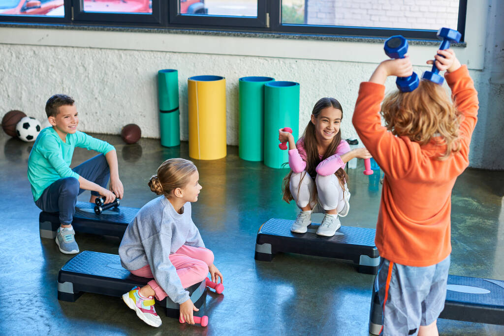 vier fröhliche Kinder posieren auf Fitnessstiefeln mit Hanteln und lächeln fröhlich, Kindersport - Foto, Bild
