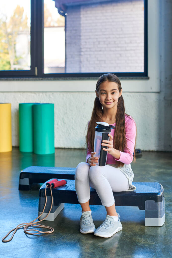vertikale Aufnahme des kleinen Smiley-Mädchens auf Fitness-Stepper sitzend mit Wasserflasche, Kindersport - Foto, Bild