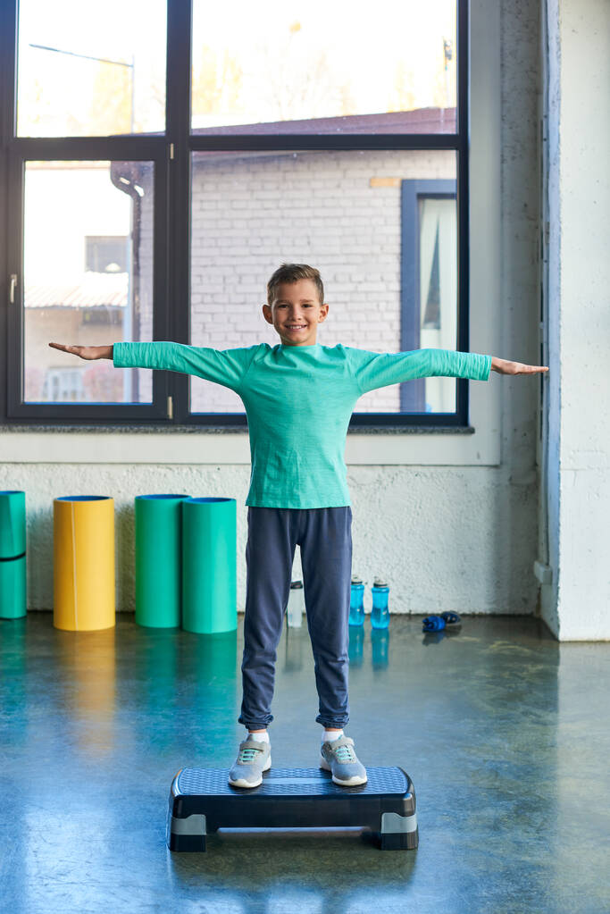 счастливый маленький симпатичный мальчик, стоящий на фитнес-стопе и радостно улыбающийся на камеру, детский спорт - Фото, изображение