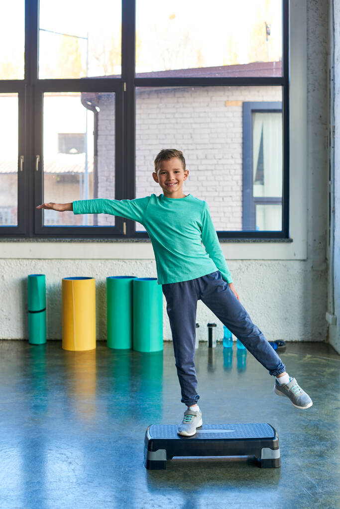 счастливый маленький мальчик растягивается на фитнес-ступенька и весело улыбается на камеру, детский спорт - Фото, изображение