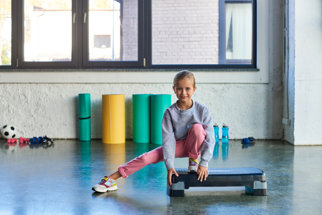 Маленькая симпатичная девочка в спортивной одежде, растягивающая ногу на фитнес-ступеньку, глядя в камеру, детский спорт - Фото, изображение