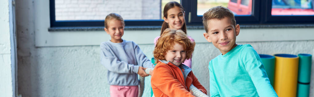 χαρούμενα παιδιά σε αθλητικά ρούχα τραβώντας σχοινί γυμναστικής στο γυμναστήριο και χαμογελώντας χαρούμενα, παιδικά σπορ, πανό - Φωτογραφία, εικόνα