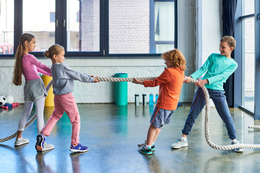 preadolescent children in sportswear playing tug of war with fitness rope in gym, child sport - Photo, Image