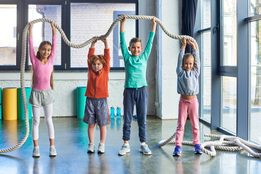 bambini allegri in abbigliamento sportivo alzando la corda fitness sopra la testa e sorridendo alla macchina fotografica, sport infantile - Foto, immagini