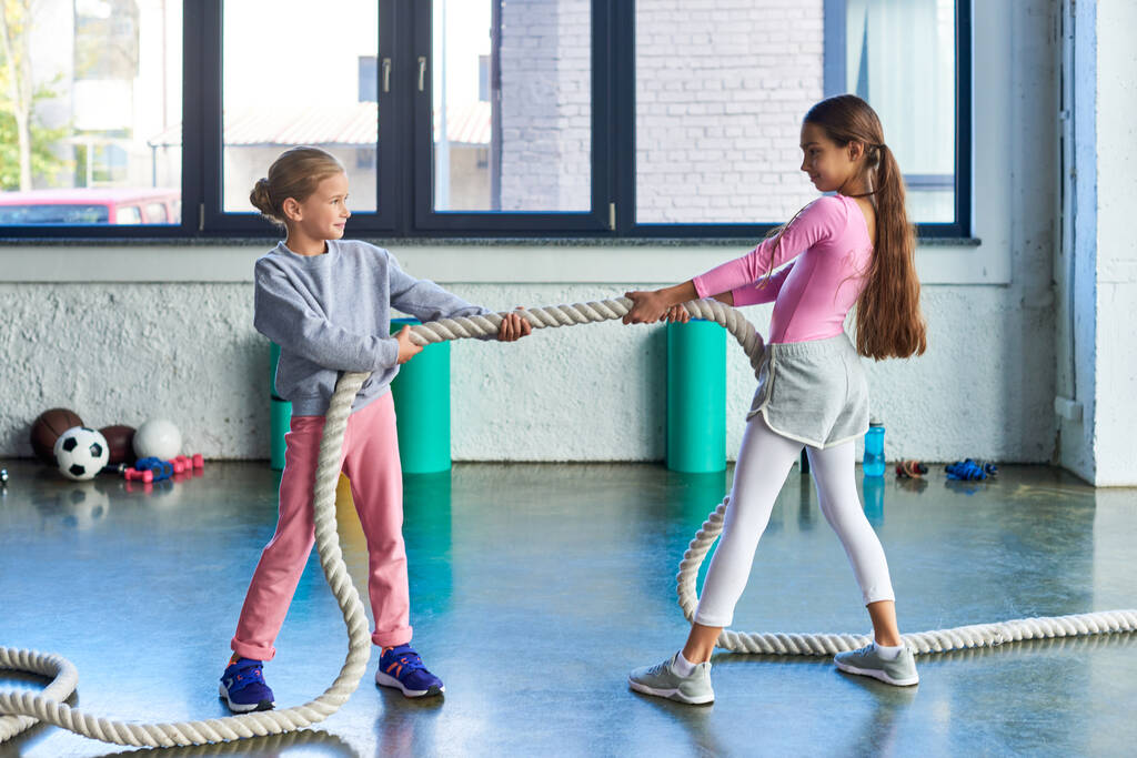 cheerful little girls in sportswear pulling fitness rope and smiling joyfully at each other, sport - Photo, Image