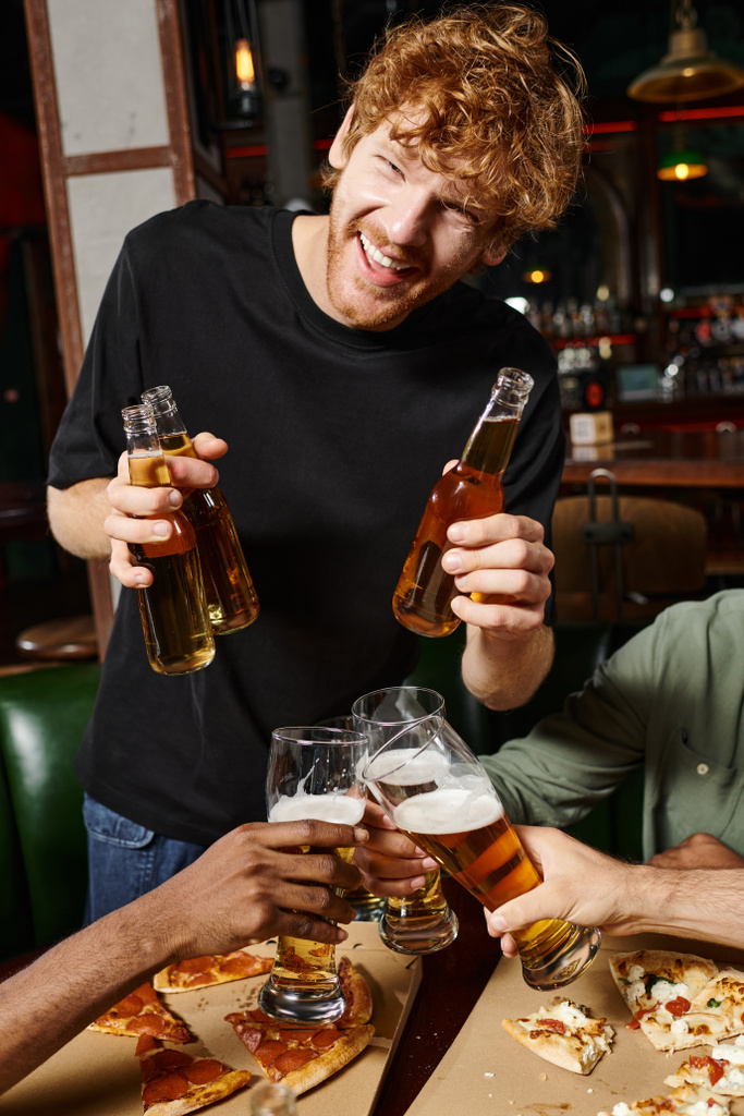 glücklicher rothaariger Mann mit lockigem Haar, der Flaschen mit Bier in der Nähe von Freunden hält, die Gläser in der Bar prosten - Foto, Bild