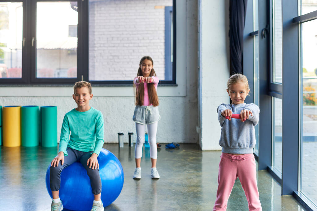 schattig jongetje zitten op fitness bal met twee mooie meisjes oefenen met halters, kind sport - Foto, afbeelding