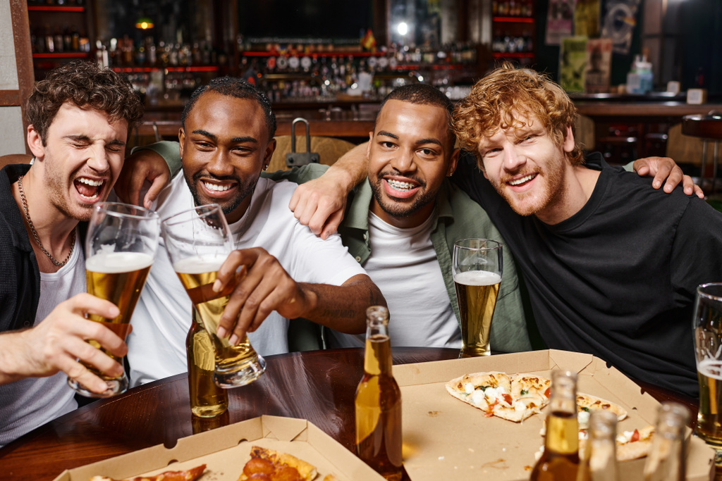 gruppo di amici multietnici felici che si abbracciano e brindano con bicchieri di birra, trascorrendo del tempo al bar - Foto, immagini
