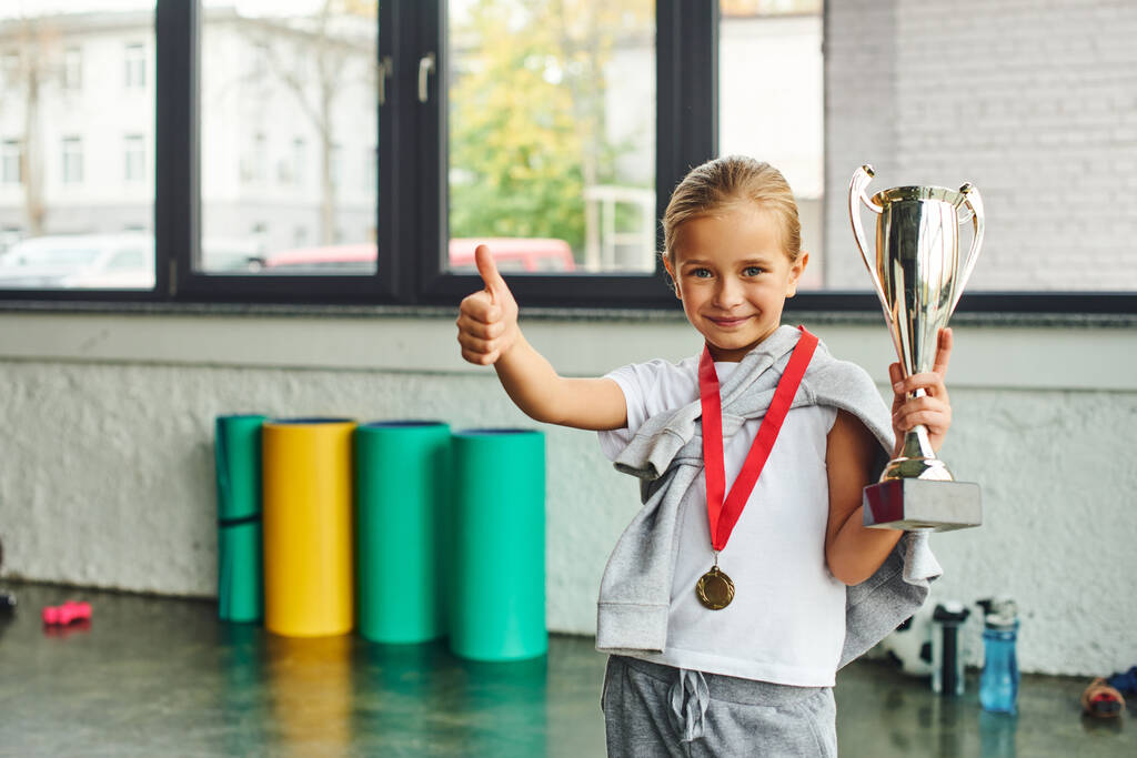 malá blondýnka dívka pózující s trofejí a zlatou medailí a ukazující palec nahoru na kameru, dětský sport - Fotografie, Obrázek