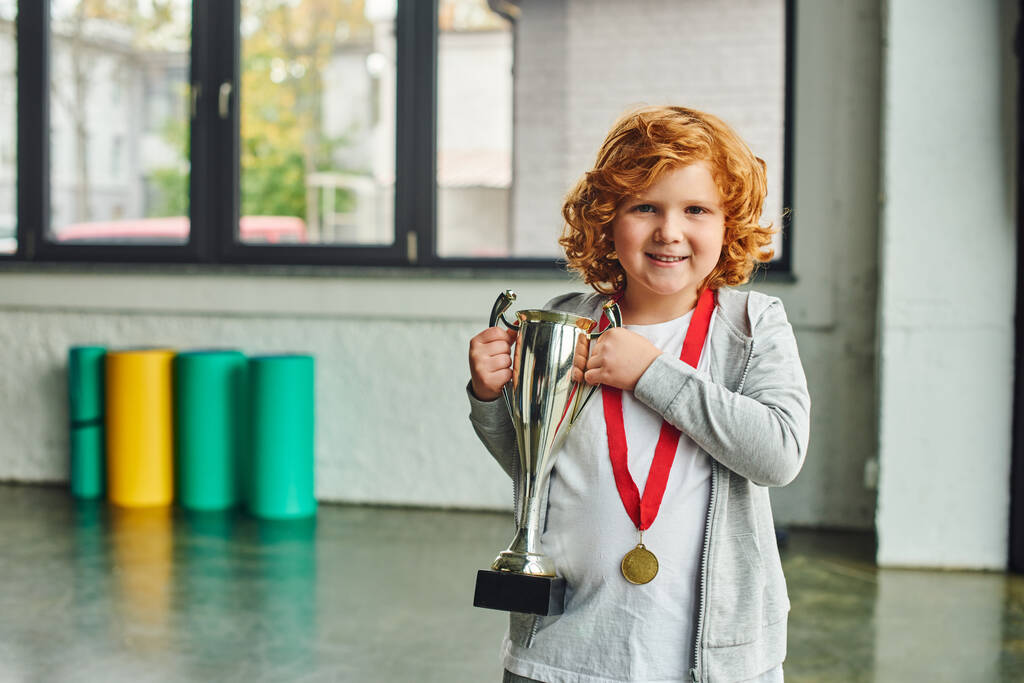 pěkný červenovlasý chlapec se zlatou medailí drží obrovskou trofej a vesele se usmívá na kameru - Fotografie, Obrázek