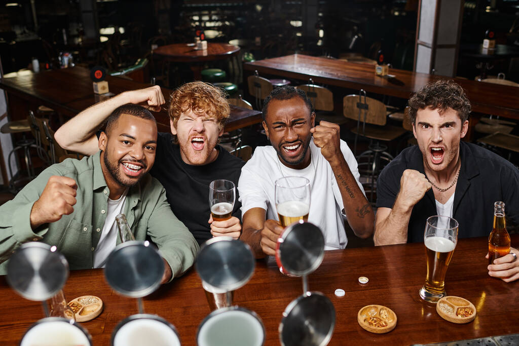 quatro excitados fãs de futebol multiétnico segurando copos de cerveja e torcida, amigos do sexo masculino no bar - Foto, Imagem