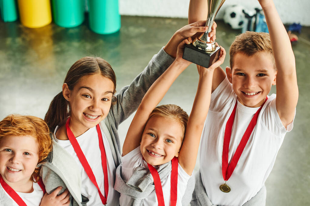 vier fröhliche, süße vorpubertäre Kinder in Sportkleidung triumphieren mit riesiger Trophäe in der Hand - Foto, Bild