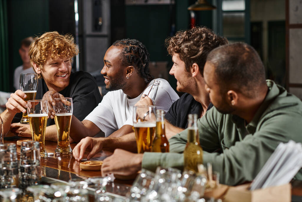 Мультикультурные мужчины болтают и пьют пиво, счастливые друзья проводят время вместе в баре - Фото, изображение