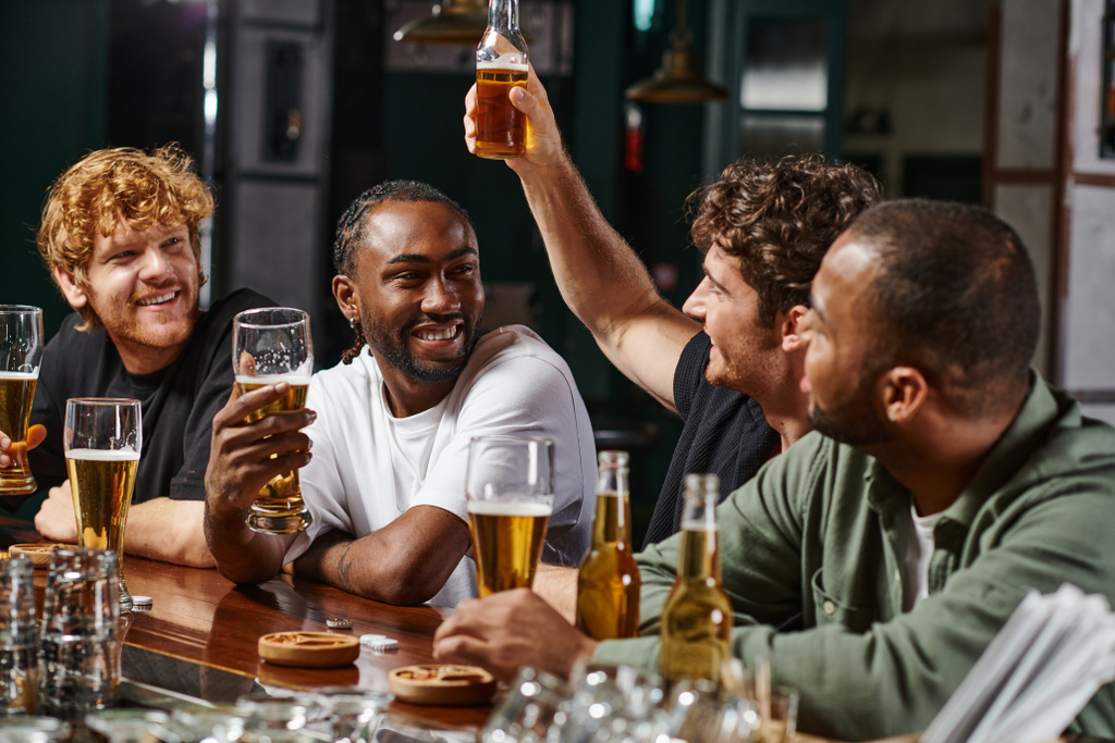 счастливые мультикультурные мужчины смотрят на друга, поднимающего бокал пива, проводя время вместе в баре - Фото, изображение