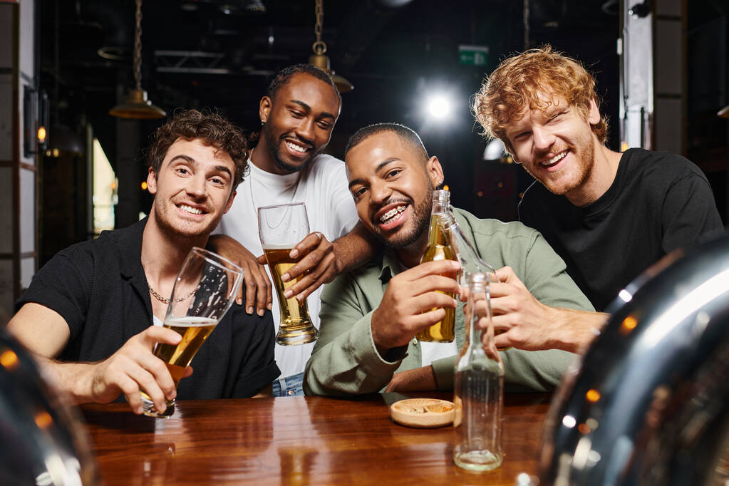 czterech szczęśliwych wielokulturowych mężczyzn patrzących w kamerę trzymając okulary z piwem w barze, męscy przyjaciele - Zdjęcie, obraz
