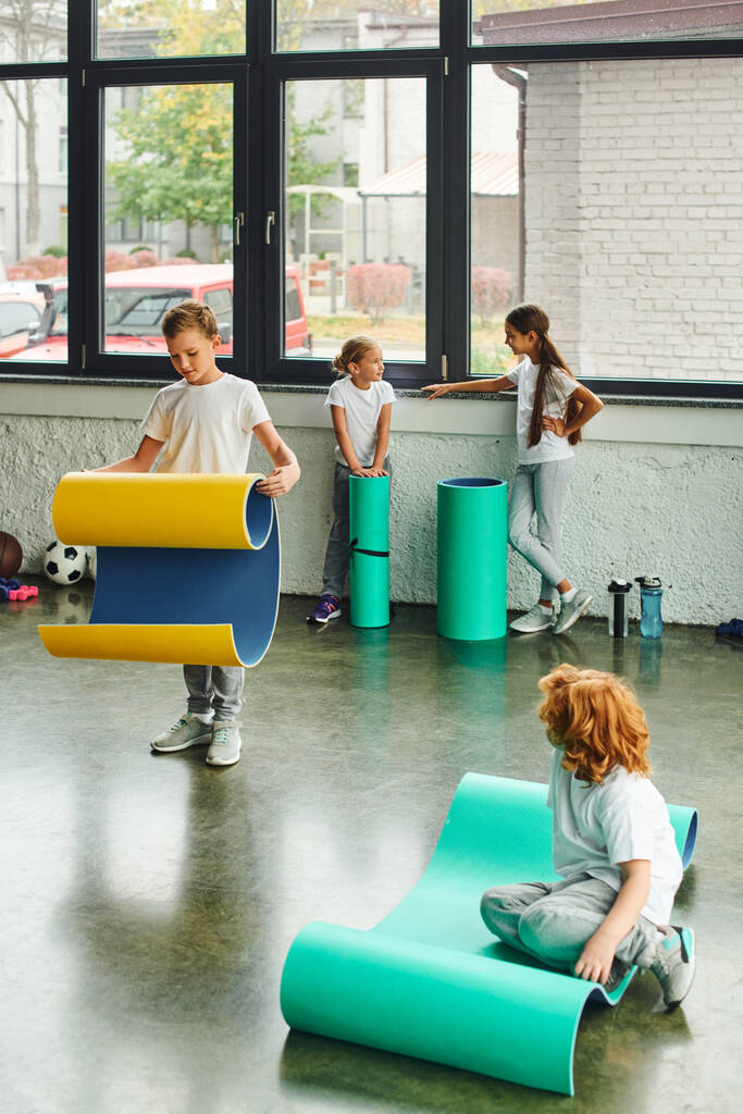 two little boys unfolding fitness mats while girls talking to each other on backdrop, child sport - Photo, Image