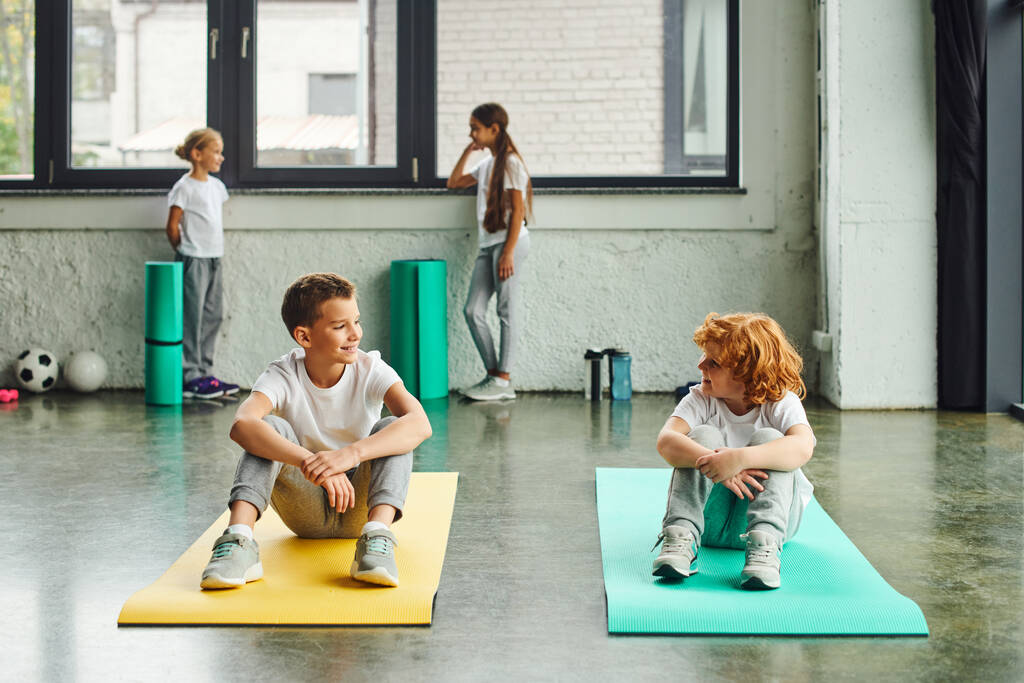twee vrolijke jongens glimlachen naar elkaar op fitness matten met leuke meisjes staan op de achtergrond - Foto, afbeelding