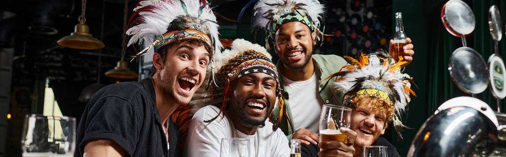 spandoek, multi-etnische mannelijke vrienden in hoofddeksel met veren toasten bier en tijd doorbrengen in bar - Foto, afbeelding