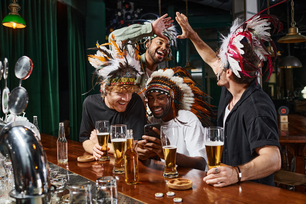 щасливі мультикультурні чоловіки в головній убір з пір'ям, які спостерігають за футбольним матчем на смартфоні в барі - Фото, зображення