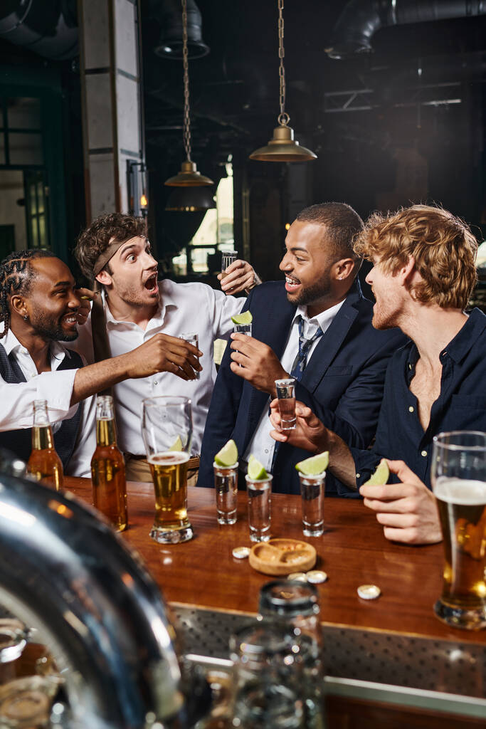 fröhliche interrassische Freunde halten Tequila-Shots mit Limette in der Nähe von Biergläsern auf dem Tresen - Foto, Bild