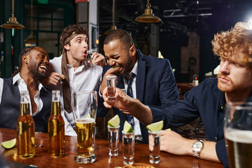 candid photo of funny and drunk interracial men in formal wear drinking tequila in bar after work - Photo, Image