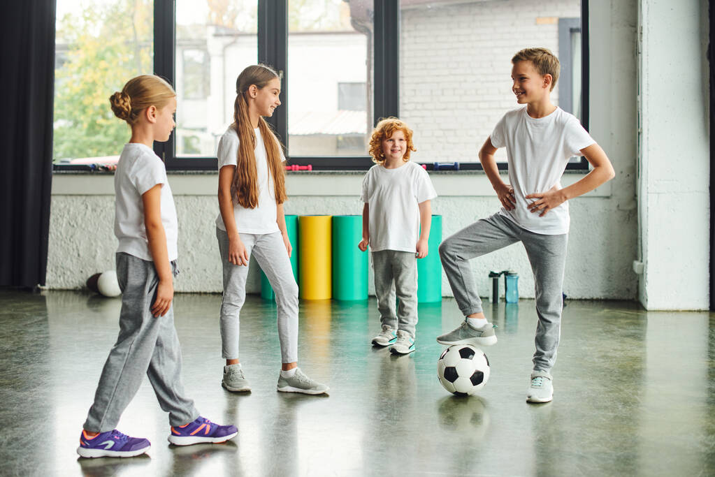 deux filles et un garçon roux souriant à un autre garçon mettant sa jambe sur le ballon de football, sport d'enfant - Photo, image